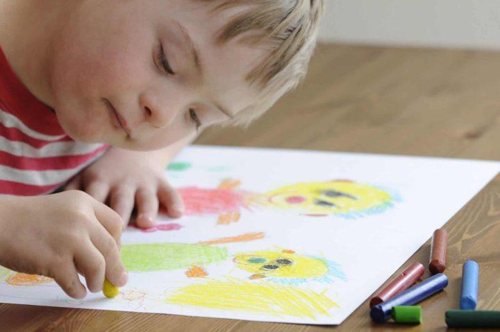 Child with Down syndrome drawing colorful figures with crayons on paper.